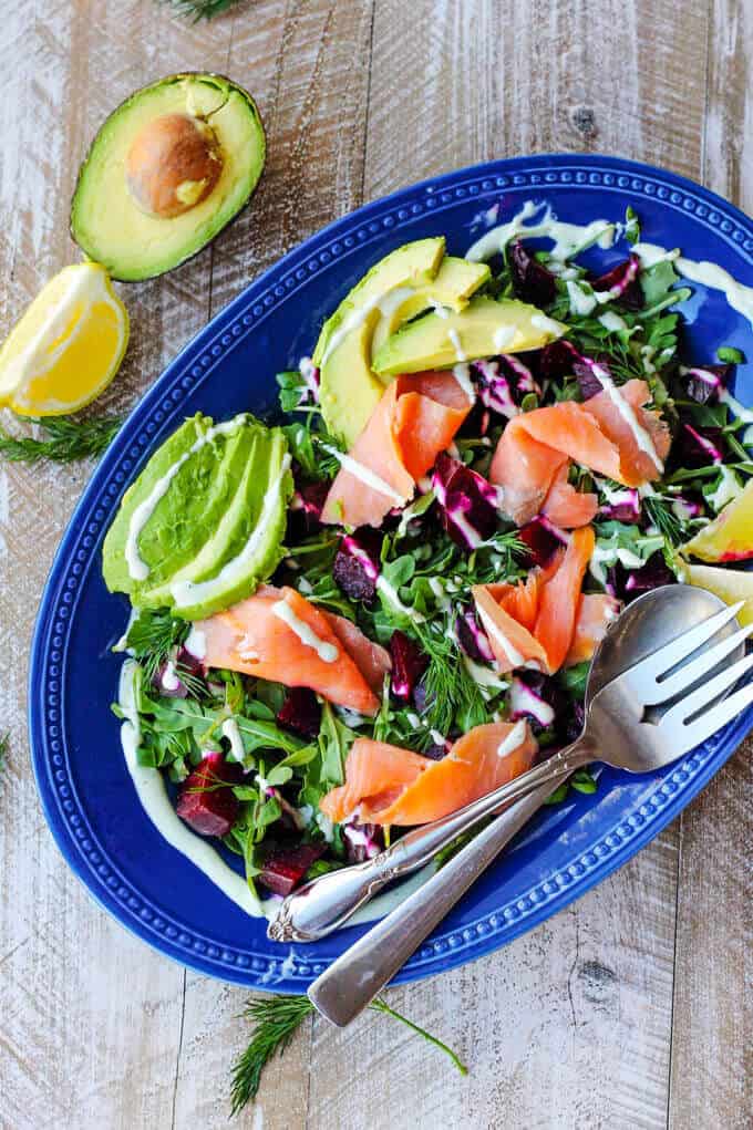 Smoked Salmon and Roasted Beets Salad with avocado, lemon quarters and a Goat Cheese Dressing on a blue pate placed diagonal with fork and spoon; with half avocado and lemon on a side