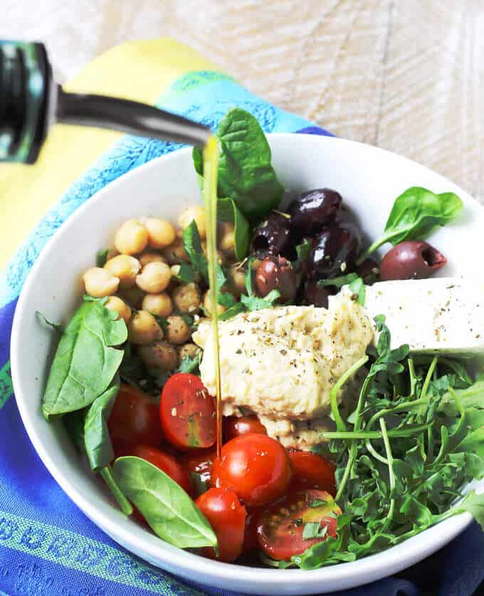 Mediterranean Hummus Bowl with feta tomatoes greens olives and chickpeas with olive oil pouring over the bowl