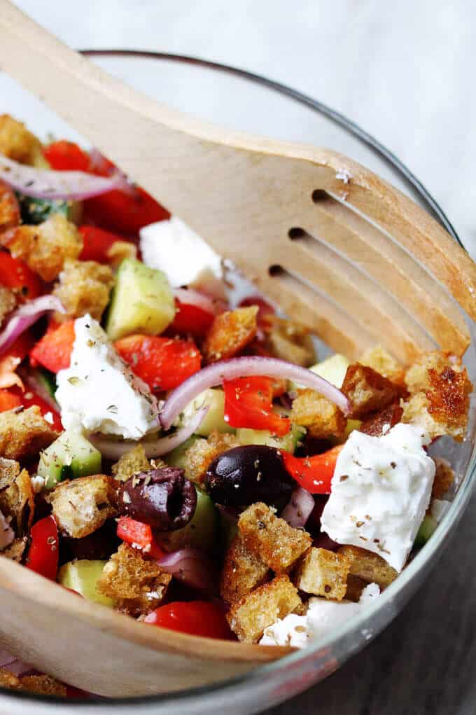 Greek Panzanella Salad in a bowl with wooden spoon and fork, combines traditional Greek salad flavors, like Kalamata olives, and tangy feta with bread cubes toasted in tasty garlicky vinaigrette.