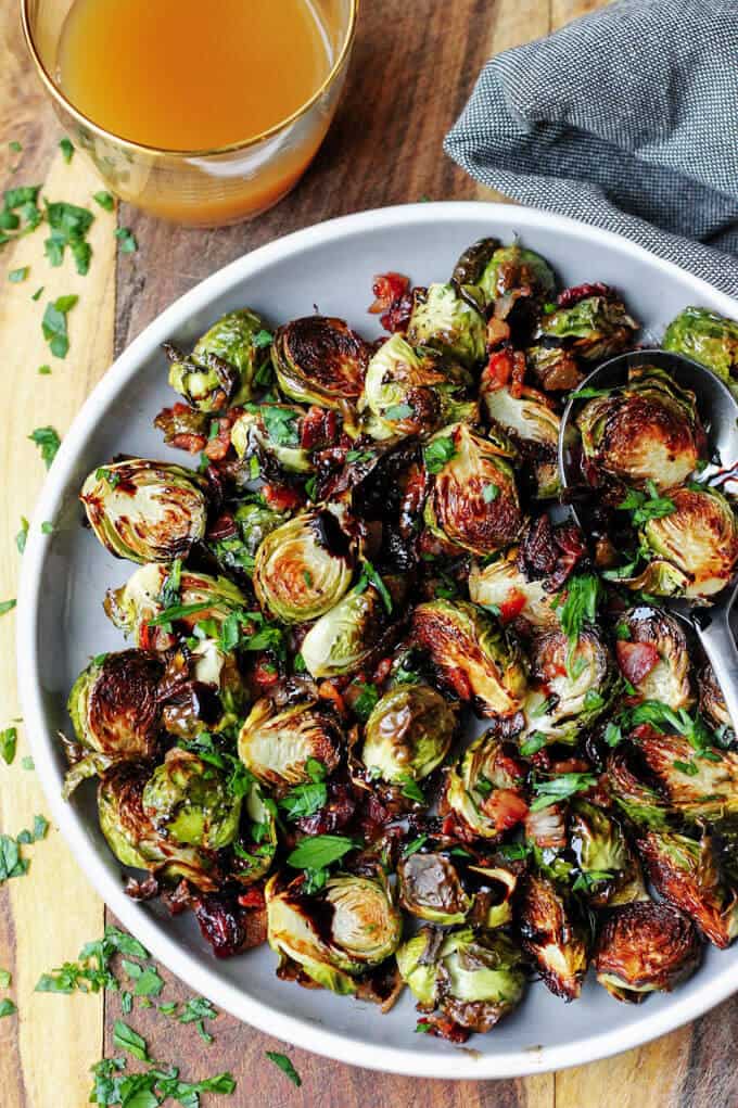 Balsamic Glazed Brussels Sprouts with Bacon on a grey plate with drink and kitchen towel on a side