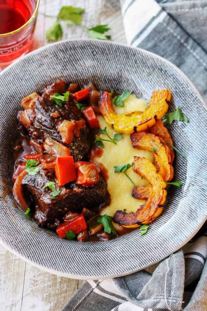 Red Wine Braised Short Ribs with Parmesan Polenta and Roasted Delicata Squash in a grey plate with kitchen towel and drink on a side