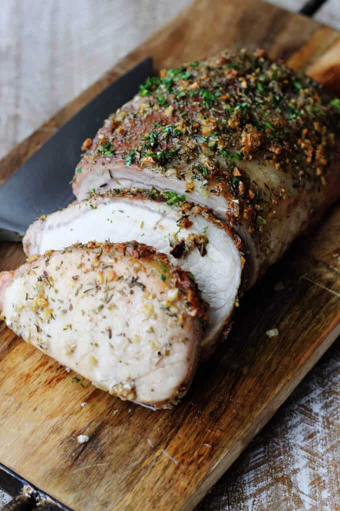 Brown sugar pork loin with garlic and herbs on a cutting board, partially cut up with knife on a side
