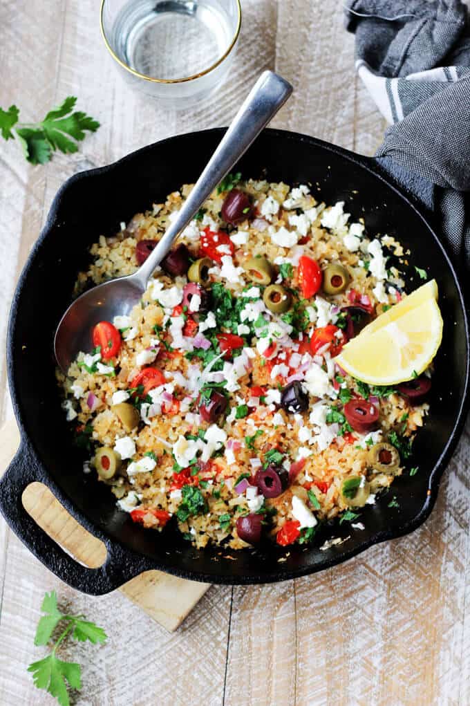 Cauliflower fried rice with tomatoes, feta olives and lemon in a skillet with spoon, drink on a side