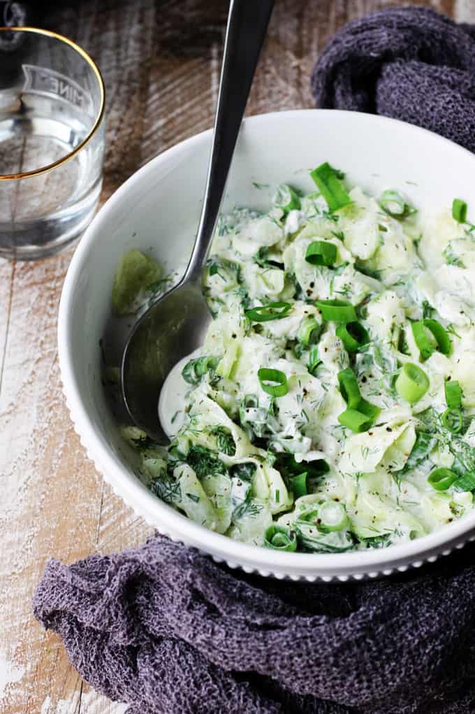 Polish cucumber salad in a white bowl with spoon in and towel around; water in a back