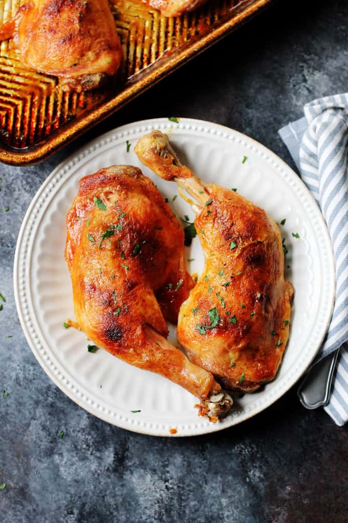 Slow roasted chicken leg quarters on a white plate with sheet pan in the back