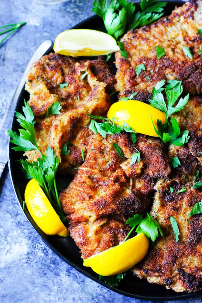 Breaded veal cutlets on a black plate with lemon pieces